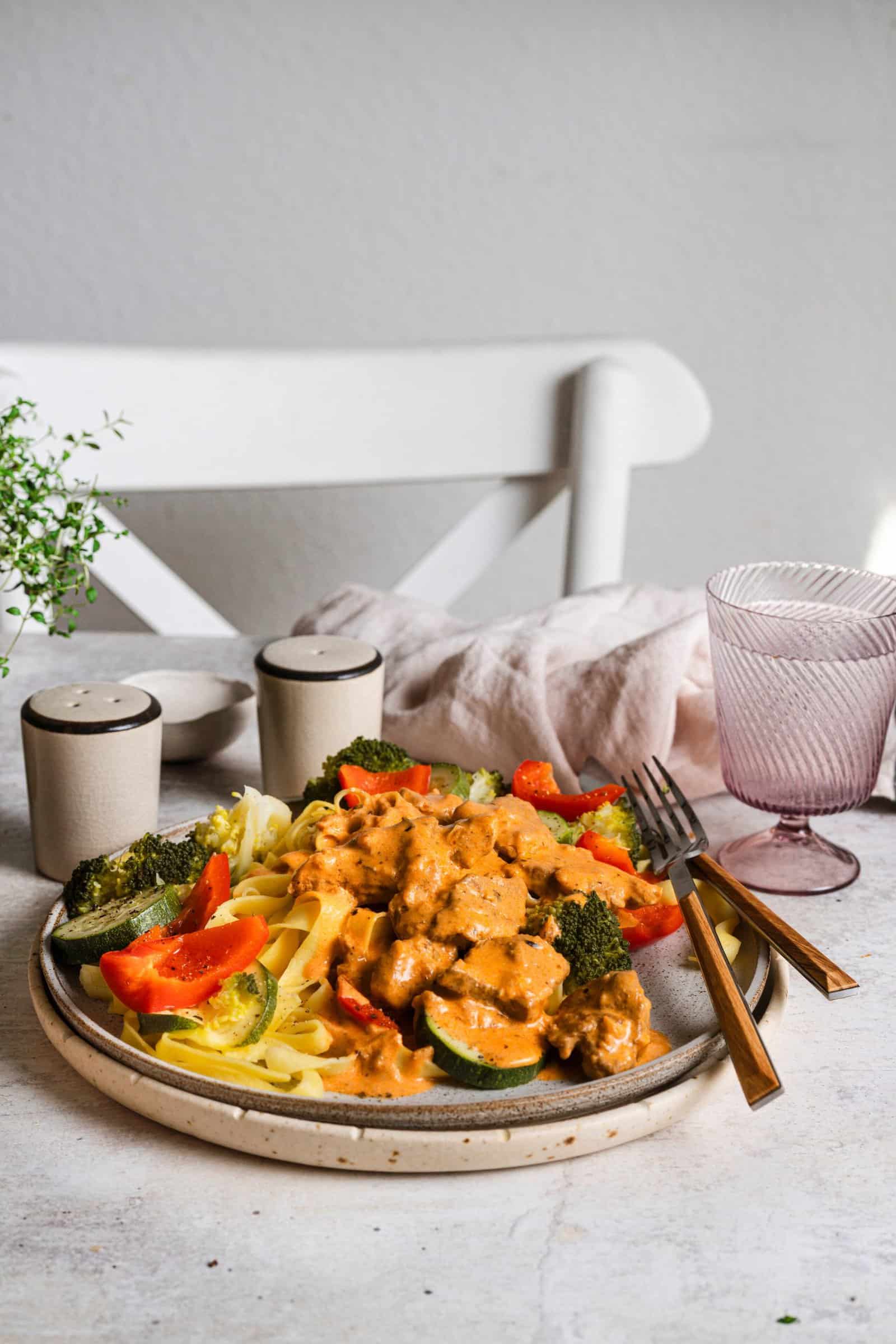 Ein Teller mit Gemüse, Pasta und Fleisch auf einem Tisch mit weißer Tischdecke, daneben Salz, Pfeffer und ein Glas
