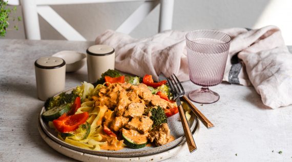 Ein Teller mit Gemüse, Pasta und Fleisch auf einem Tisch mit weißer Tischdecke, daneben Salz, Pfeffer und ein Glas