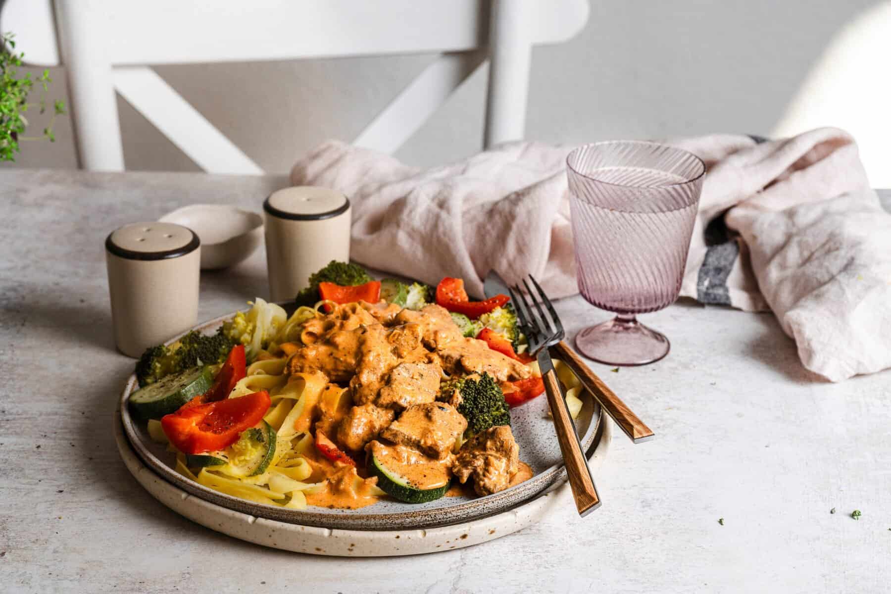 Ein Teller mit Gemüse, Pasta und Fleisch auf einem Tisch mit weißer Tischdecke, daneben Salz, Pfeffer und ein Glas
