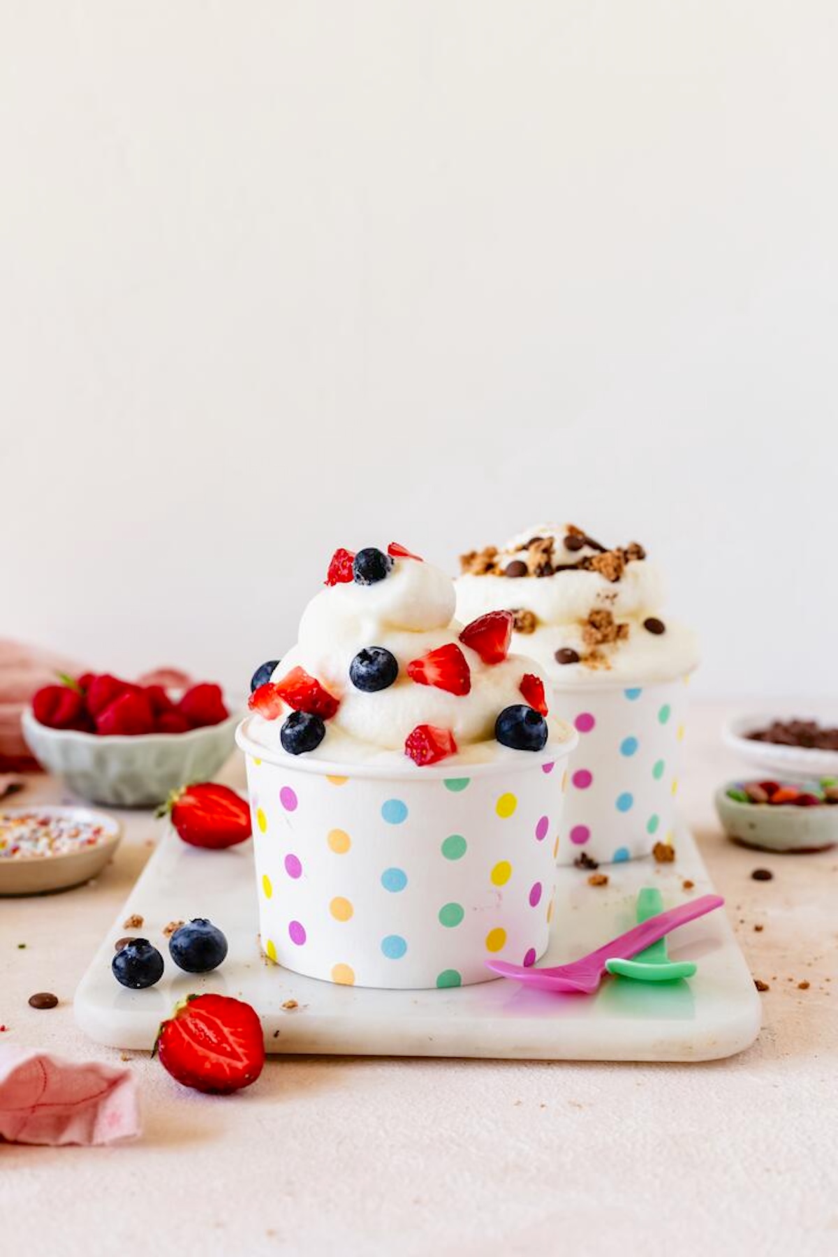 Zwei Pappbecher mit bunten Punkten auf einer Marmorplatte, darin Frozen Joghrt mit unterschiedlichen Toppings
