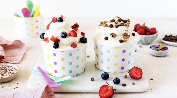 Zwei Pappbecher mit bunten Punkten auf einer Marmorplatte, darin Frozen Joghrt mit unterschiedlichen Toppings