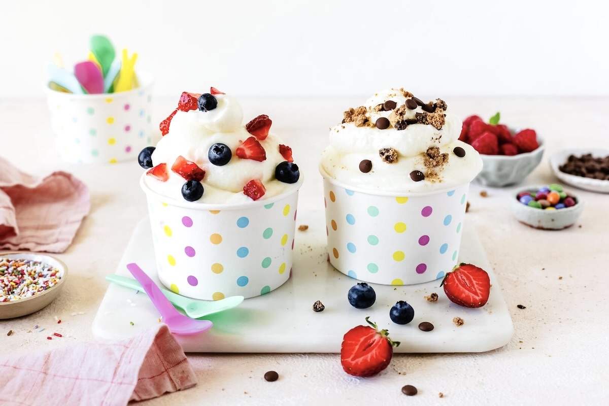 Zwei Pappbecher mit bunten Punkten auf einer Marmorplatte, darin Frozen Joghrt mit unterschiedlichen Toppings