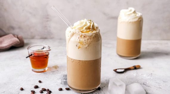 zwei Gläser mit einem Kaffeegetränk mit Sahnehaube, daneben Kaffee-Bohnen und Eiswürfel