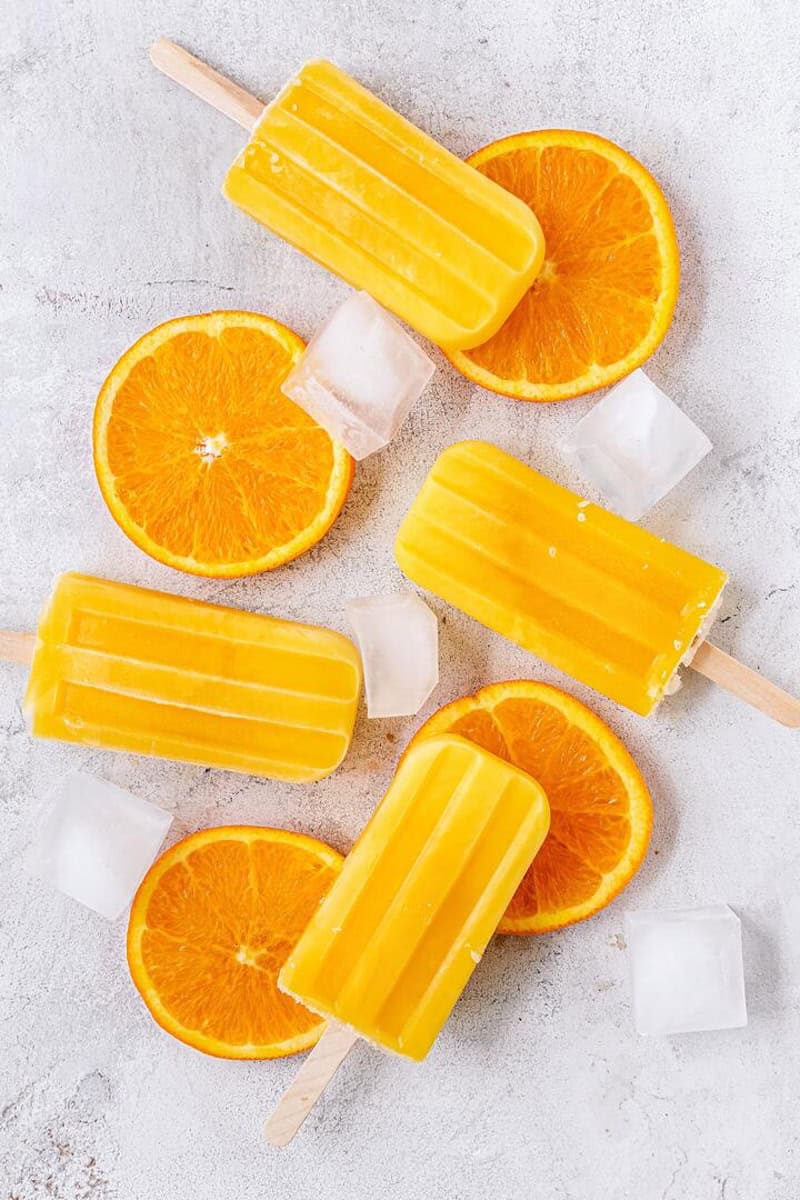Mehrere Eis am Stiel in orange, daneben Orangen-Scheiben und Eiswürfel, von oben fotografiert auf hellem Untergrund