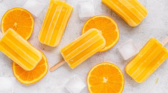 Mehrere Eis am Stiel in orange, daneben Orangen-Scheiben und Eiswürfel, von oben fotografiert auf hellem Untergrund