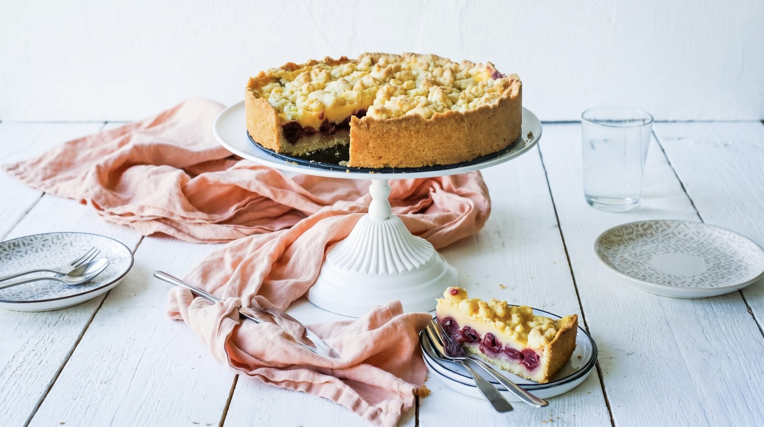 Ein Kirschkuchen auf einem Tortenständer, daneben ein Teller mit einem Stück des Kuchens, heller Tisch