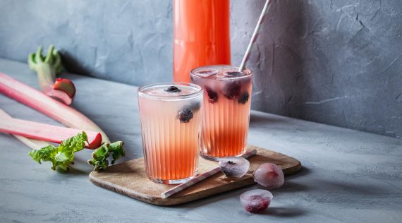 Eine Flasche mit Rhabarbersirup, zwei Gläser mit Rhabarberschorle, Rhabarber und gefrorene Früchte vor grauem Hintergrund