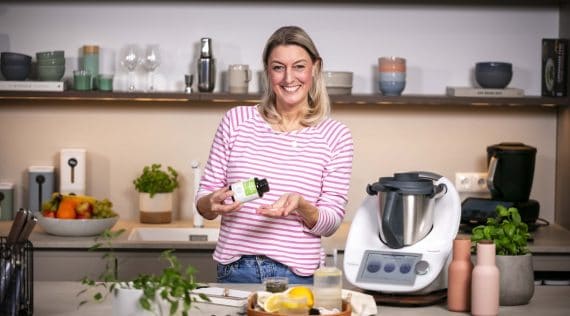 Blonde Frau in einer Küche. In der Hand ein Fläschchen mit Eisen & Vitamin C