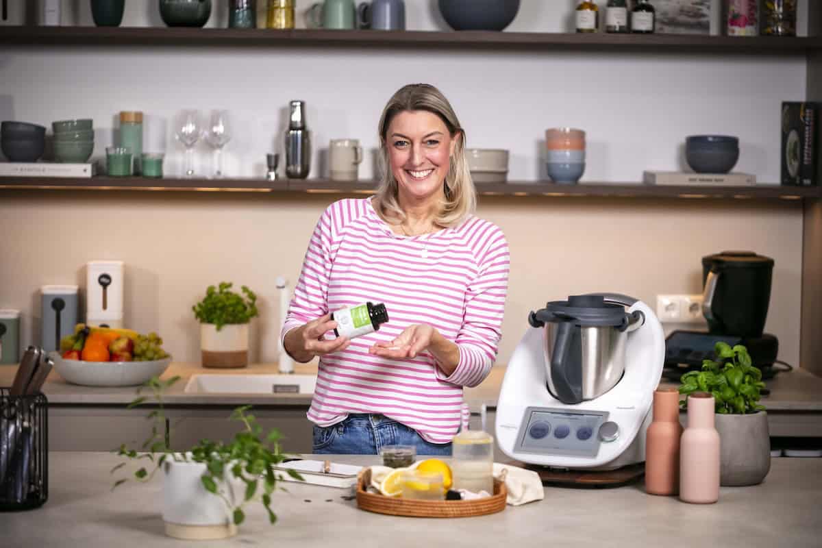 Blonde Frau in einer Küche. In der Hand ein Fläschchen mit Eisen & Vitamin C